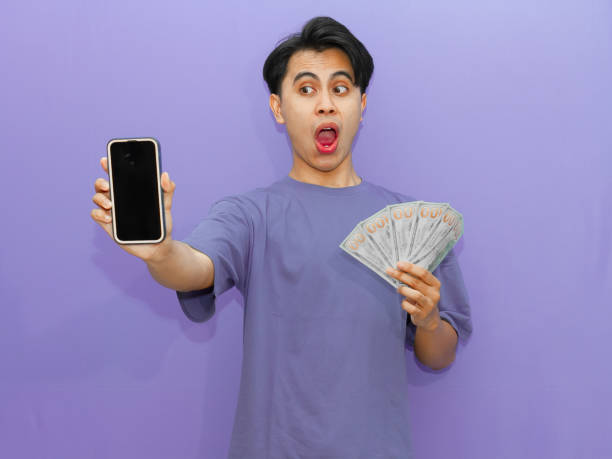 istock A young Asian man with a funny shocked expression holds a blank mobile phone screen and banknotes against a vibrant purple background. 2182731061