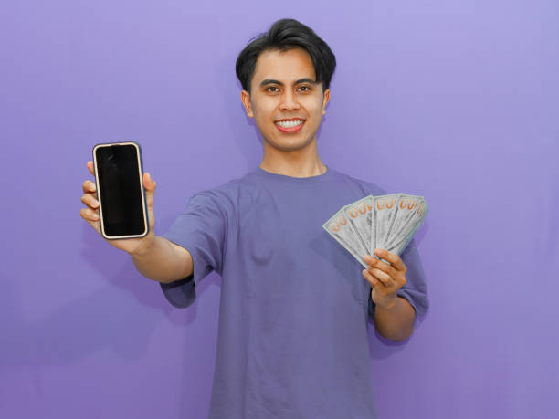 istock A smiling young Asian man displays a blank mobile phone screen while holding banknotes, set against a vibrant purple background. 2182731063