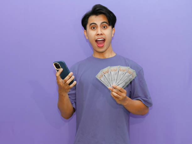 istock Young Asian man with surprised expression, holding money and using mobile phone, isolated on purple background. Perfect for advertising and promotional use. 2182732789