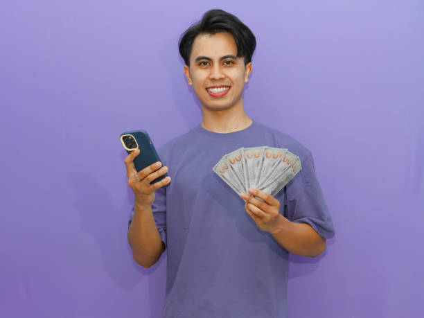 Young Asian man smiling confidently while holding a smartphone and money on a purple background, perfect for advertising, finance, and success themes.