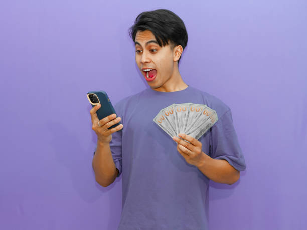 istock Young Asian man with surprised expression, holding money and using mobile phone, isolated on purple background. Perfect for advertising and promotional use. 2182732851