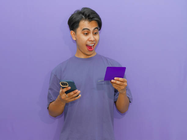 istock Young Asian man holding a savings book and mobile phone with an amazed expression, standing against a purple background. 2182736576