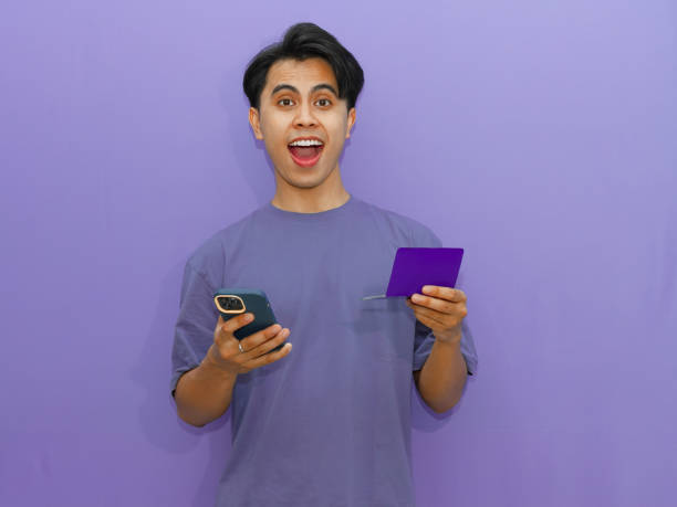 istock Young Asian man holding a savings book and mobile phone with an amazed expression, standing against a purple background. 2182736580