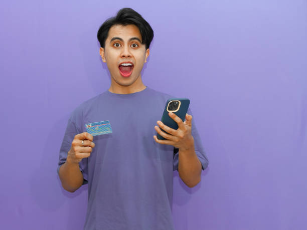 istock Young Asian man surprised while holding his resident identity card (KTP) and using a cellphone against a purple background. 2182737360