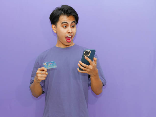 istock Young Asian man surprised while holding his resident identity card (KTP) and using a cellphone against a purple background. 2182737525
