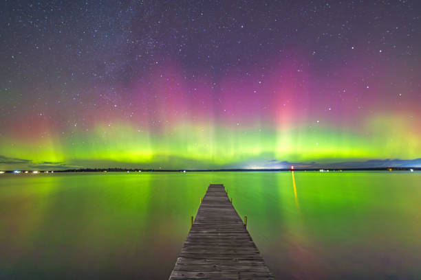 Vibrant colors of green and pink auroras dance across the night sky, mirrored beautifully on a calm lake. A wooden pier extends into the water, leading the eye towards the horizon filled with shimmering stars. Higgins Lake, Michigan. 11/8/24