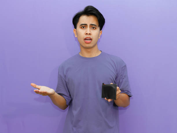 istock Poor young Asian man with an empty wallet, displaying a sad and shocked expression against an isolated purple background. 2182739306