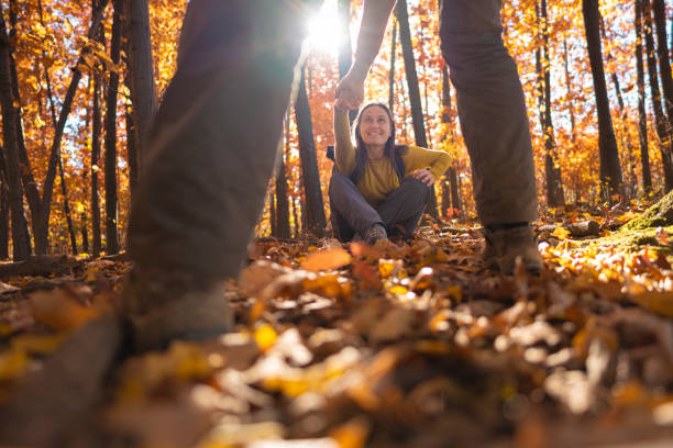 Man gives hand to his girlfriend to stand up from fall yellow leaves carpet and go hiking in autumn forest. Happy mature couple sharing an adventure together.