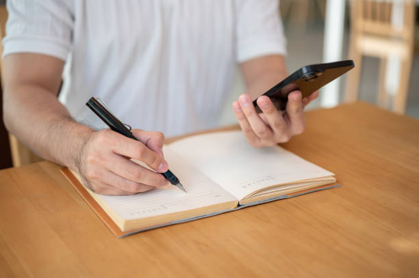 istock A cropped shot of an Asian man holding his smartphone while taking notes in his notebook. 2182769025