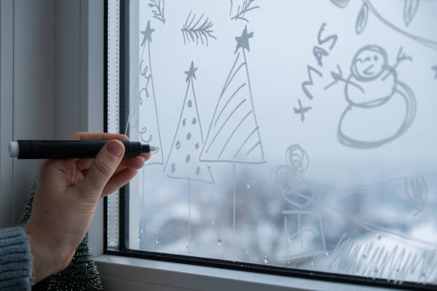 istock Creative enthusiastic unrecognizable woman in sweater paint window with white marker. Christmas tree decoration on big clear window. Snowy winter outside 2182778991