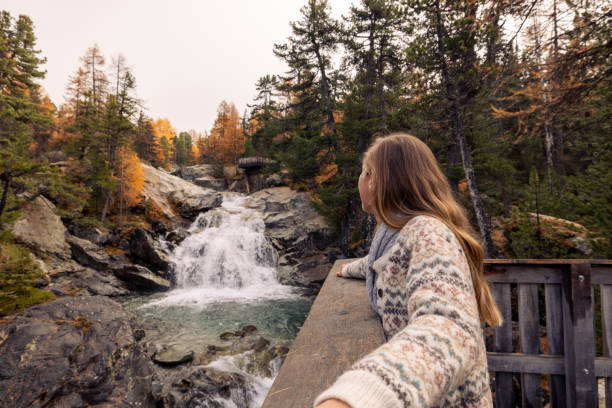 Woman in cozy sweater enjoys the view of a serene waterfall in an autumn forest, embracing nature's beauty on a peaceful day