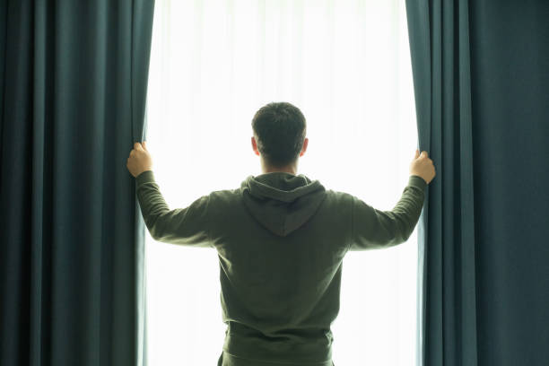 istock Young man looks out window and opens the curtains, rear view. 2182783326