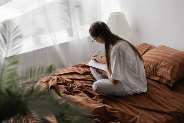 istock lifestyle image of young woman sitting on her bed and making notes to her diary. 2182787721