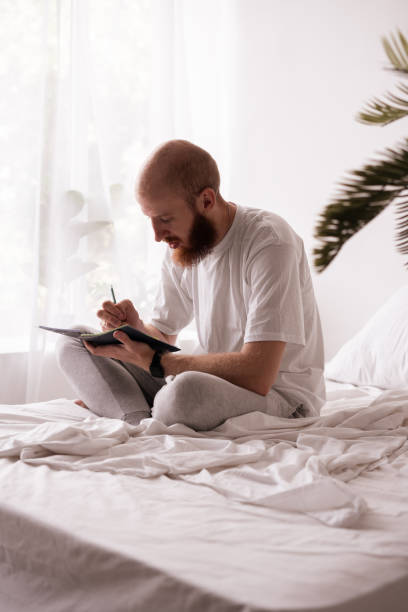 istock Young handsome man writing a letter or notes in the diary sitting on the bed in his room. Successful business ideas. 2182788131