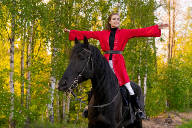 Woman is riding a black horse in a forest. She is wearing a red coat and is smiling