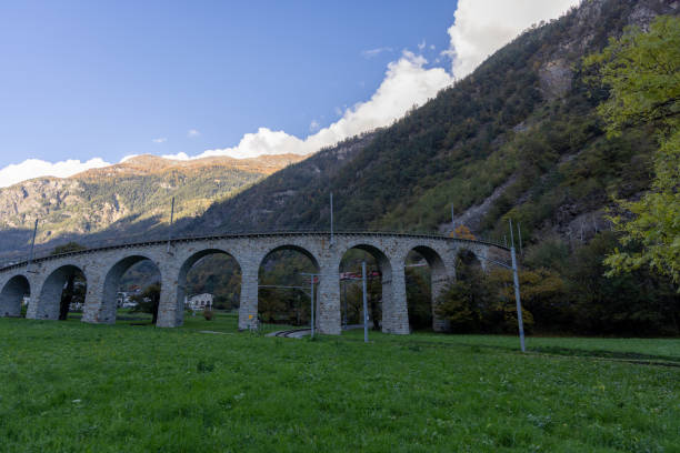 A scenic stone arch railway bridge stretches across a green valley with mountains rising in the background under a clear blue sky.