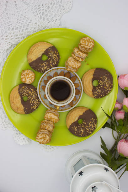 Top view of a coffee cup surrounded by almond-decorated chocolate cookies and cream-filled biscuits on a bright green plate. Pink flowers and a white lantern add warmth to the setup.