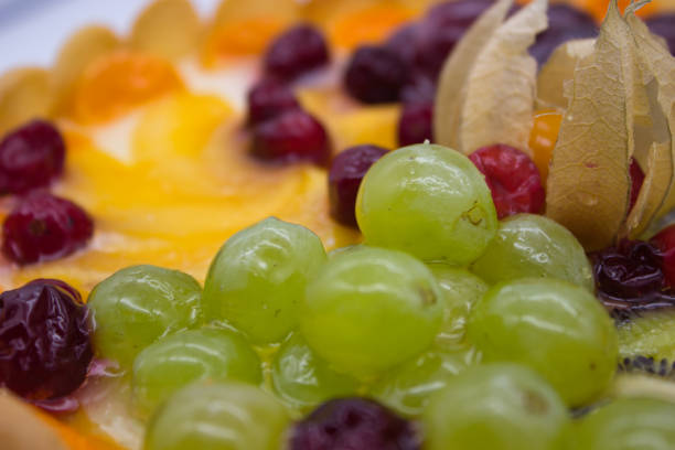 This close-up of a vibrant fruit tart highlights an array of juicy berries, grapes, kiwi, and physalis arranged on a creamy filling. The glaze adds a fresh, glossy texture that enhances the visual appeal of the dessert.