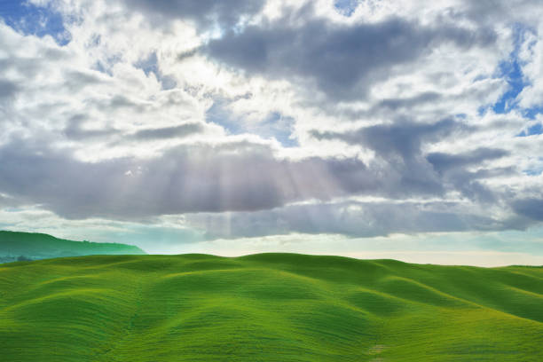 Sun rays pierce through dramatic clouds, casting light over lush, green rolling hills in a serene rural landscape. The contrast between the vibrant hills and moody sky highlights the natural beauty of the Tuscan countryside.