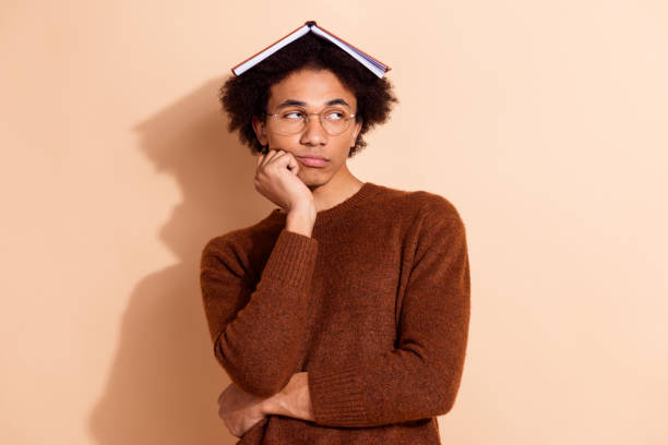 istock Young African American student with a book on his head looking thoughtful against beige background 2182801721