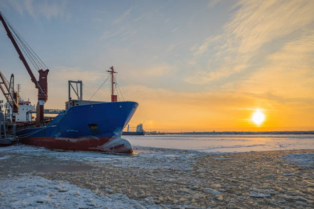 In winter, in severe frost, a cargo ship stands on a pier for loading against the background of a frozen river and the setting sun