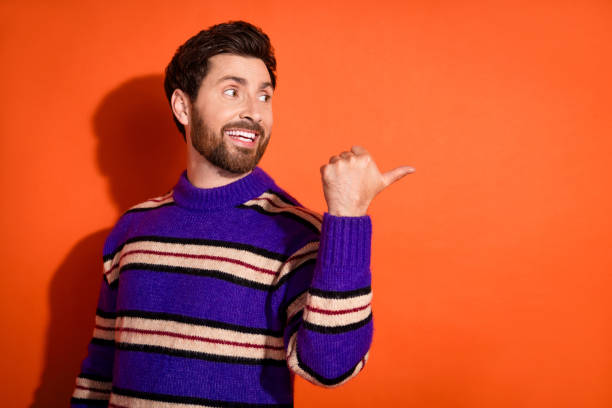 istock Cheerful young man with a beard in a striped blue sweater pointing back over an orange background, embodying confidence and positivity 2182809724