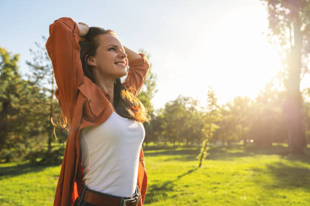 optimistic caucasian young woman enjoying springtime outdoors walking in city park. woman stands with hands behind head and smiling. - ontgiften fotos stockfoto's en -beelden