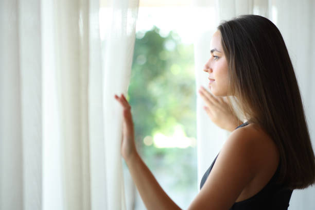 istock Serious woman looking through a window opening curtain 2182823614