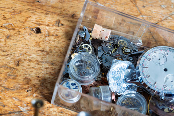 Collection of delicate watch parts, tools, and small gears housed in a transparent box, resting on a rustic wooden workbench, indicating a workshop setting for fine, intricate assembly work