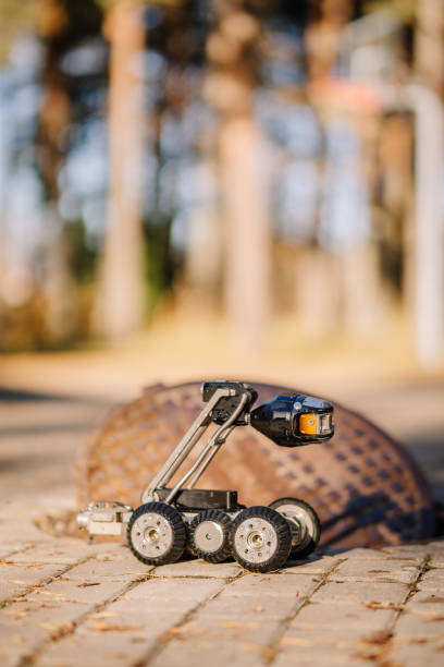 A four-wheeled inspection robot with a camera stands on a paved surface near an open manhole, used for utility and sewer inspection.