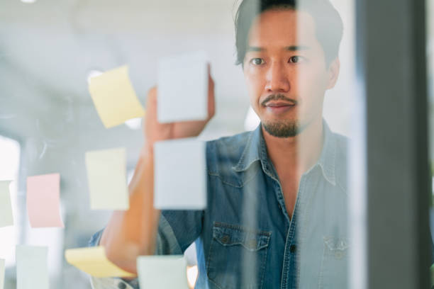 istock Focused on his task, man in denim shirt is writing on sticky notes attached to glass board, showcasing collaborative work environment. His concentration reflects importance of teamwork and creativity 2182850181