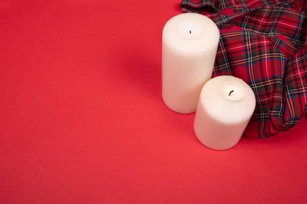 Two white candles placed on a vibrant red background next to a red plaid fabric, creating a cozy and warm atmosphere. Perfect for seasonal, interior design, or holiday-themed visuals that emphasize warmth and simplicity.