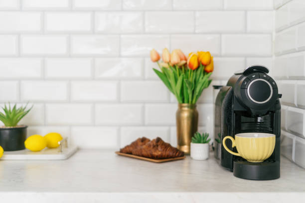 A bright kitchen showcases a coffee maker next to a yellow cup with espresso, vibrant flowers, fresh lemons, and a croissant, perfect for a relaxing breakfast moment