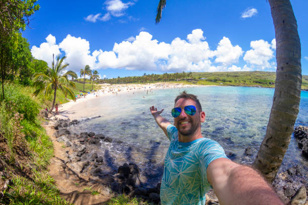 A handsome young happiness cheerful joyful fit latin male backpacker tourist visiting Anakena Beach Easter Island - Rapa Nui, in the middle of Pacific Ocean, part of Polynesia / Chile in Latin America; posing for a picture in front of the Moai's sculptures with his open outstretched arms at Anakena Beach, wearing a blue t-shirt and blue stylish sunglasses, in a sunny summer hot day. Man visiting and sightseeing Latin America destination. Latin America / Chile travel concept.