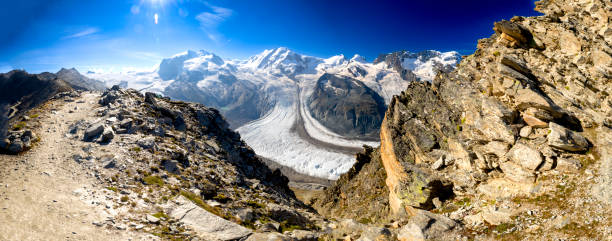 Zermatt, Switzerland - A rugged mountain trail from Gornergrat to Hohtalli offers stunning panoramic views of the Gorner Glacier and surrounding alpine peaks under a clear blue sky.