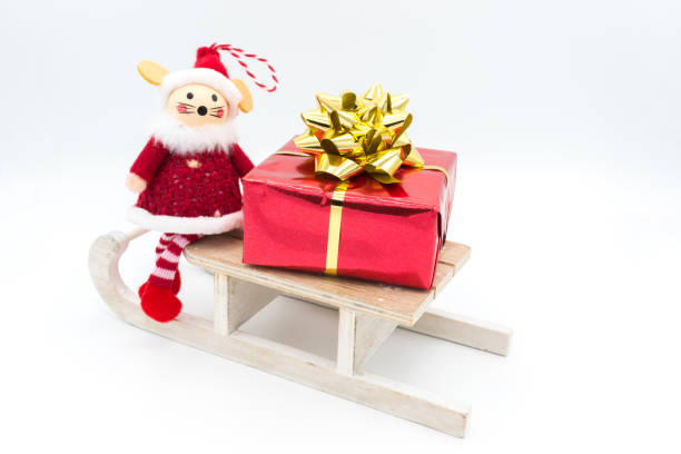 Red gift package with a golden ribbon, placed on a wooden sledge with a Christmas toy mouse decoration. Isolated on a white background.