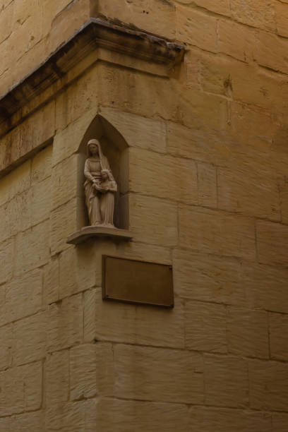 A serene stone statue of the Virgin Mary adorns an old sandstone wall corner, reflecting religious tranquility and historical architecture under soft lighting. Ideal for themes of spirituality and heritage.