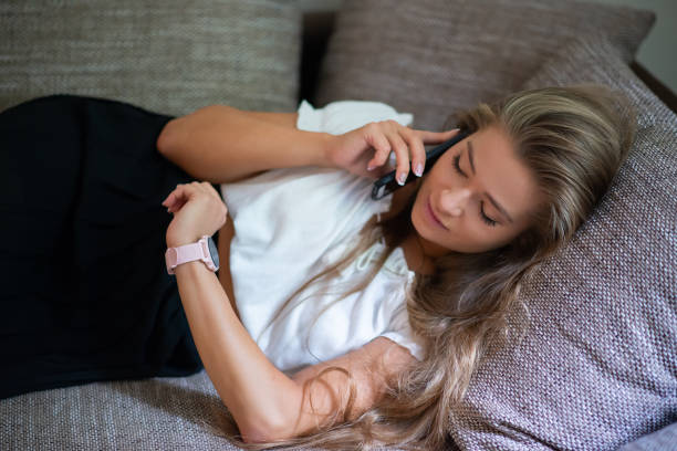 istock Young woman relaxing on sofa while talking on phone and checking smartwatch 2182903152