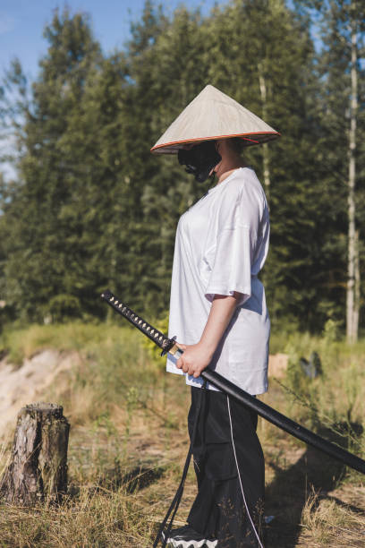 istock The cosplayer holds a katana while wearing a conical hat, white shirt and black pants in a forest setting, creating a dramatic and intriguing scene. The samurai trains with a katana on the beach. 2182905451