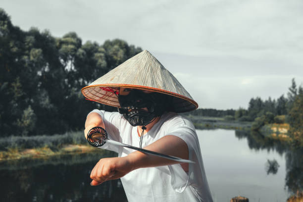 istock A samurai warrior wearing a conical hat and a black mask unsheathes a katana by a river, demonstrating traditional Japanese martial arts and warrior culture. The warrior practices with a katana. 2182906482