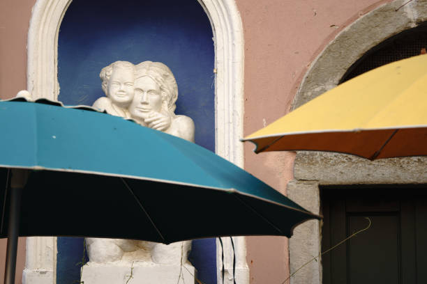 Manarola, Italy - November 2, 2024: A stone relief sculpture depicting a mother and child is set against a deep blue background within an arched recess on a building's facade. The artwork is partially framed by two vibrant umbrellas, one teal and one yellow, creating a contrast of colors against the pastel wall.