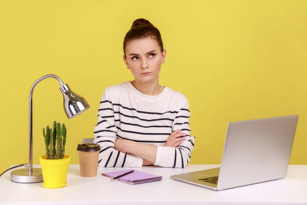 istock Woman in striped shirt sitting with thoughtful confused expression while working on laptop in office 2182925983