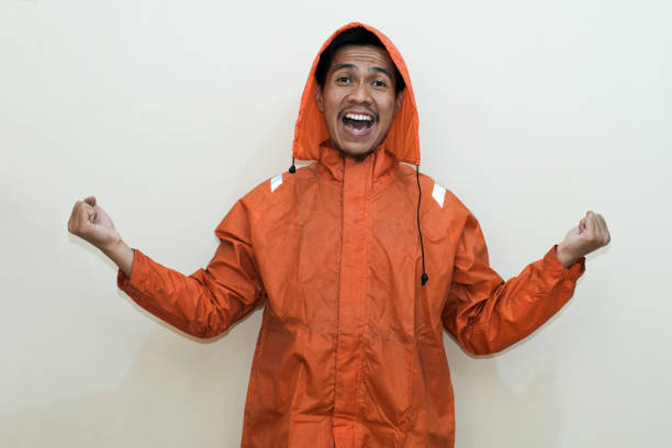 Exciting Southeast Asian man wearing orange raincoat with hand shows winning victory gesture. Positive person. standing over isolated white background. rainy season.