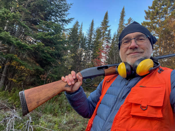 Proud mature man with his shotgun and ear protection ready to hunt in the forest during day of autumn.