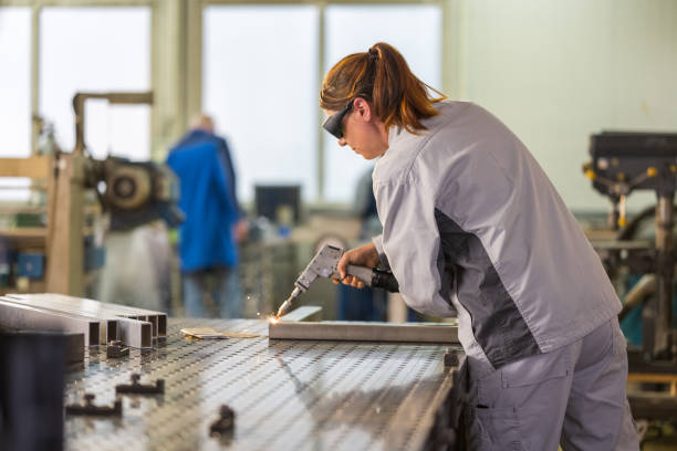 A young adult Caucasian woman engages in precise welding on an aluminum structure, clad in protective work gear, in an industrial setting.