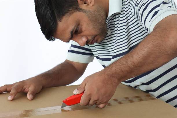 Stock photo of man is seen carefully opening a cardboard delivery parcel with a sharp knife, slicing through the packaging to reveal its contents. Focused and precise, he handles the parcel with caution, showcasing the process of unpacking a delivery