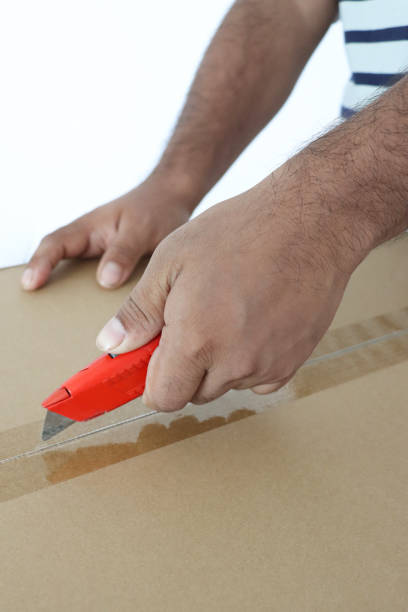 Stock photo of a man carefully opens a cardboard delivery box with a sharp knife, slicing through the packaging with precision. His hands are steady as he handles the parcel, reflecting the anticipation of unboxing. This close-up image captures the moment of unpacking a delivery