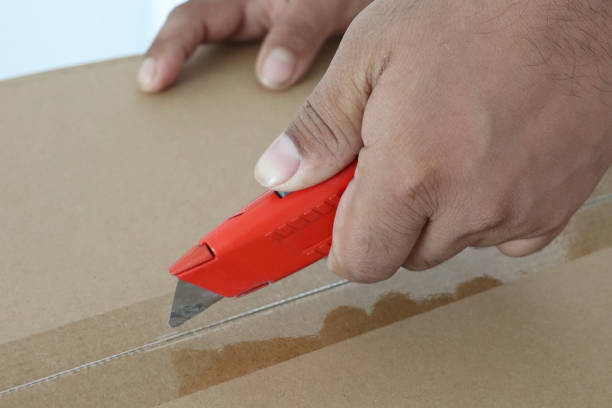 Stock photo of a man's hands are seen carefully opening a cardboard delivery parcel with a sharp knife. The precise handling of the knife as he cuts through the tape captures the anticipation and care in unboxing a new package