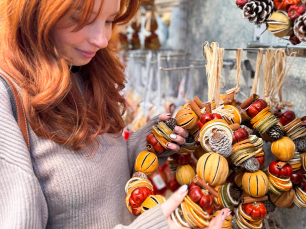 Stock photo showing a close-up view of Christmas potpourri wreath decorations hanging on a retail display, by raffia loop, made from wire frame strung with whole split and sliced dried citrus fruits (oranges and limes), pine cones, mini pumpkins and cinnamon sticks. imagen de fotograma completo de una atractiva mujer pelirroja que mira las decoraciones navideñas, coronas de marco de alambre ensartadas con cítricos secos enteros divididos y cortados en rodajas (naranjas y limas), piñas, mini calabazas y palitos de c