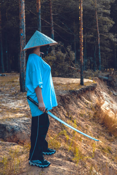 A young woman dressed as a samurai, wearing a conical hat and holding a katana, stands on a sandy cliff in the forest, creating a dramatic and evocative scene. Samurai with katana.